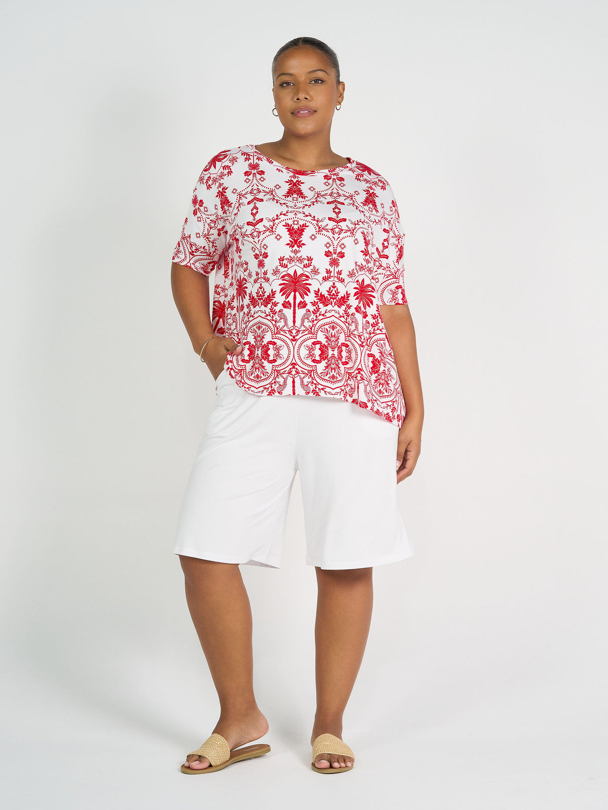 A woman stands against a white background wearing the Gina Oversized Slouch Bamboo T-Shirt in a red and white pattern, paired with white knee-length shorts, beige sandals, and hoop earrings. She poses confidently with one hand in her pocket.
