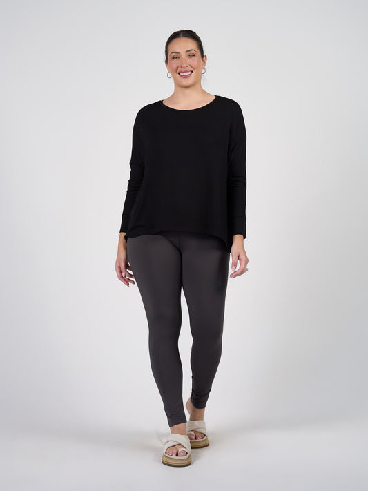 A woman smiles against a plain background, wearing the Gina Long Sleeved Bamboo Top in black, dark gray leggings, hoop earrings, and light-colored platform sandals.
