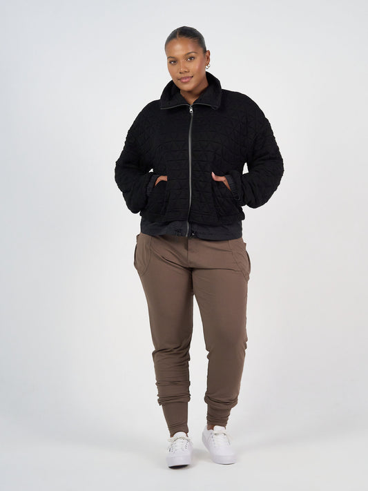 A person faces forward against a plain light background, hands in pockets, wearing the Grace Quilted Bamboo Zip Up Jacket in black, brown jogger pants, and white sneakers.