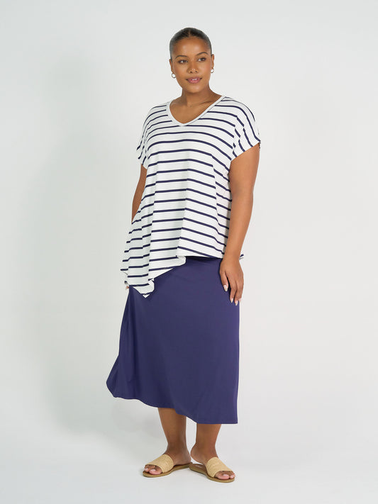 A woman stands against a white background wearing the Holiday Asymmetrical Bamboo V-Neck T-Shirt in blue & white stripe with a navy skirt and tan slides, looking slightly to the side with a relaxed expression.