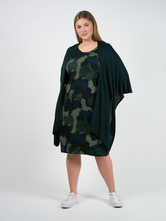 A woman with straight, light brown hair poses confidently against a white background, wearing a green and black camo dress, white sneakers, and the Supreme Cashmere/Bamboo Travel Wrap in Dark Green—a luxury travel accessory.