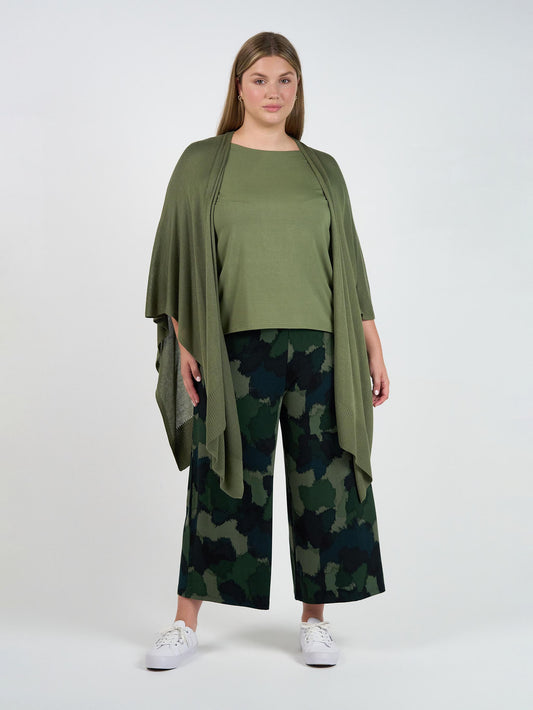 A woman with long hair and a neutral expression stands against a white background, wearing an olive green top, matching draped cardigan, white sneakers, and Ciara 7/8 Length Bamboo Culottes in camouflage print.
