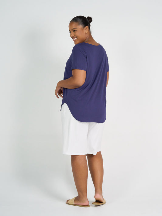 A woman smiles over her shoulder, wearing a loose blue Janis Short Sleeve Bamboo T-Shirt, white knee-length shorts, and sandals against a plain light background.