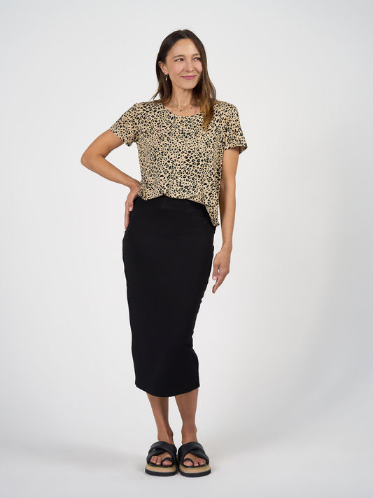 A woman stands and smiles in the Janis Short Sleeve Bamboo T-Shirt - Brown Leopard, paired with a black midi skirt and black platform sandals against a plain white background.