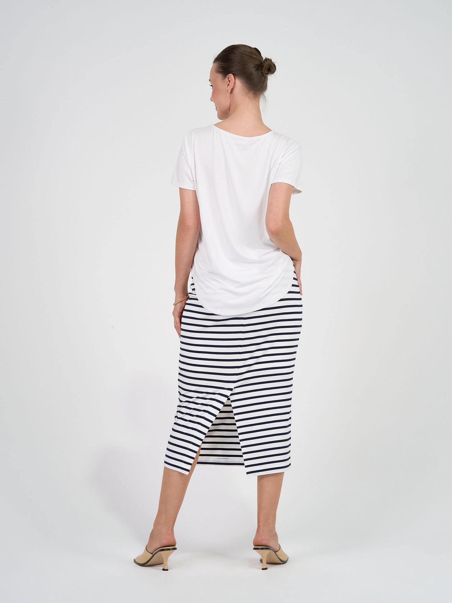 A woman with her hair in a bun poses against a white background, wearing a white short-sleeve top, the Maxi Whitney Bamboo Tube Skirt in navy and white stripe with a back slit, and beige heeled mules.