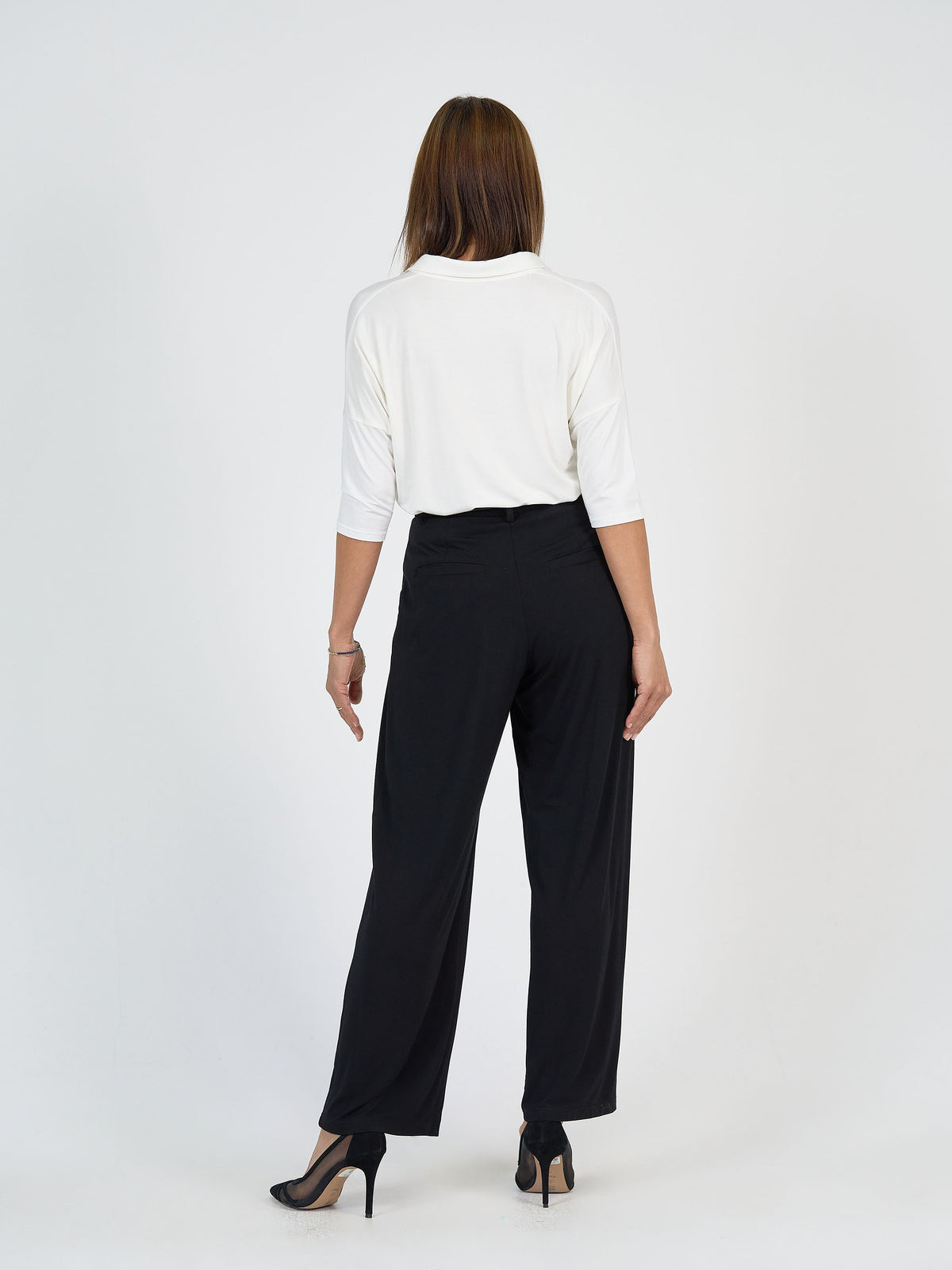 A woman stands facing away from the camera, wearing a white top with three-quarter sleeves, black high-waisted wide-leg pants, and black high-heeled shoes against a plain white background.