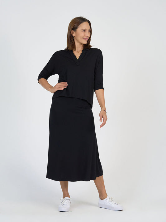 A woman stands against a plain white background, wearing a black long-sleeve top and matching black midi skirt with white sneakers. She smiles slightly and looks to her left with one hand on her hip.