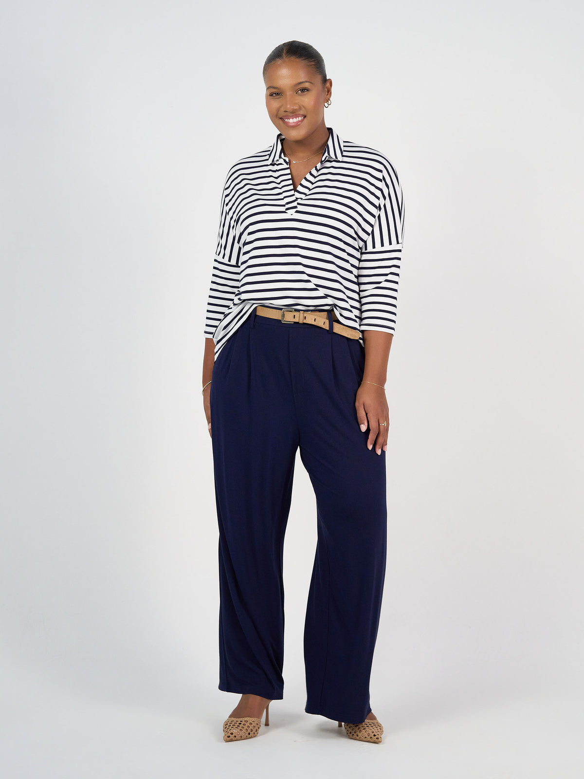 A woman with short hair smiles, wearing the Khan Bamboo Collared Polo Top in navy and white stripes tucked into high-waisted wide-leg navy pants with a beige belt and beige heels, against a plain white background.