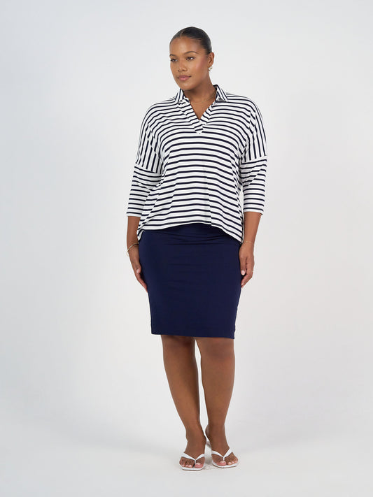 A woman stands against a plain white background wearing the Khan Bamboo Collared Polo Top in navy and white stripe, paired with a navy knee-length skirt and white sandals. Her hair is pulled back as she looks to the side.