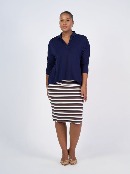 A woman stands against a plain white background wearing the Khan Bamboo Collared Polo Top in navy, a black and white striped pencil skirt, and tan pointed-toe flats.