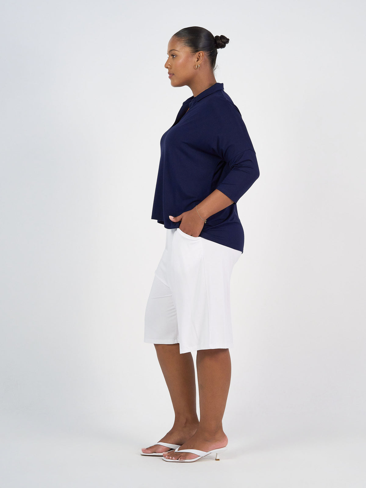 A woman stands in profile against a white background, wearing a navy blue long-sleeve top and Simone Wide Leg Bamboo Shorts in crisp white, paired with white heeled sandals. Her hair is pulled back in a neat bun and her hands are in her shorts pockets.