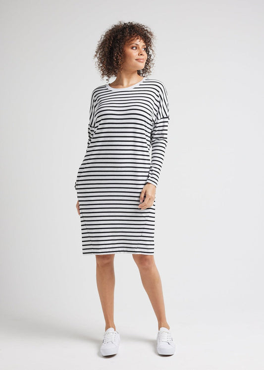 A woman stands against a plain background wearing the Turner Long-Sleeve Bamboo T-Shirt Dress in Grey Marle & Black Stripe with white sneakers, looking off to the side with a slight smile.