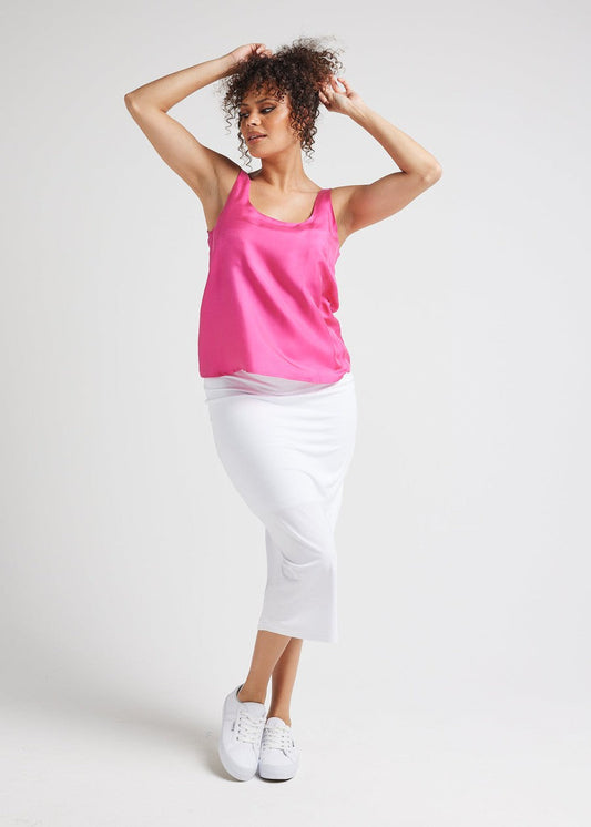 A woman with curly hair styled up poses against a white background, wearing the bright pink Poppy Silk Sleeveless Camisole paired with a fitted white midi skirt and white sneakers, her arms raised above her head.