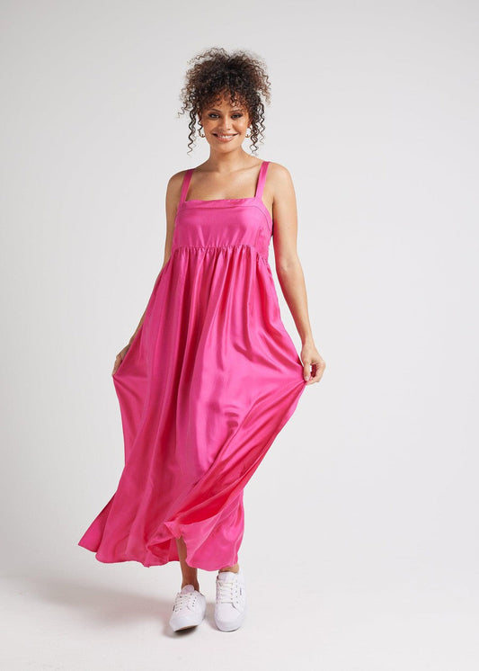 A smiling person with curly hair wears the Begonia Sleeveless Silk Maxi Dress in Fuschia and white sneakers, holding the sides of the dress against a plain light background.
