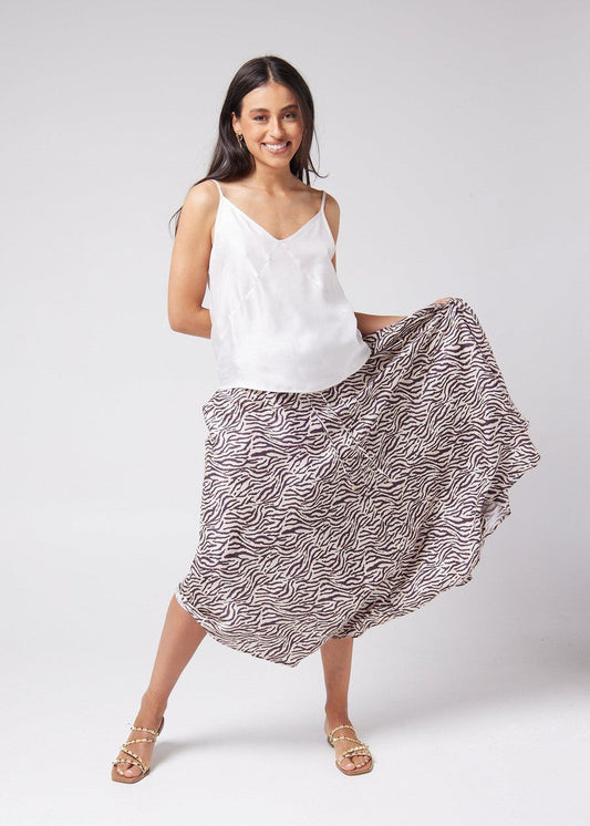 A woman smiling while holding the side of her animal printed midi skirt up and wearing white sleeveless bamboo top and a brown strap sandals