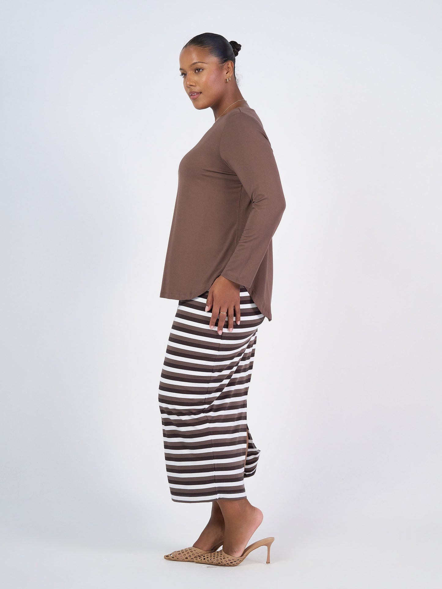 A woman stands sideways against a plain background, wearing the Maxi Bamboo Whitney Tube Skirt in brown stripe and a matching bamboo long-sleeve top, paired with beige heeled sandals. Her hair is pulled back in a neat bun.