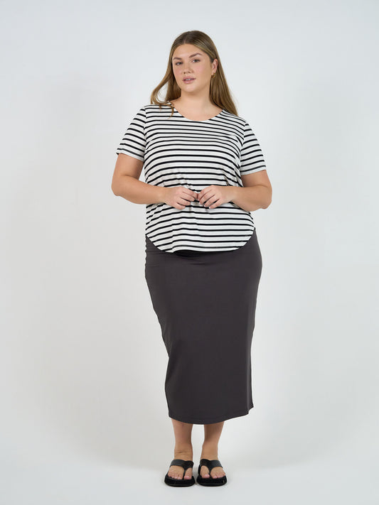 A woman with long straight hair and a neutral expression stands against a white background, wearing a black-and-white striped short-sleeve shirt, black sandals, and the Maxi Bamboo Whitney Tube Skirt in Steel.
