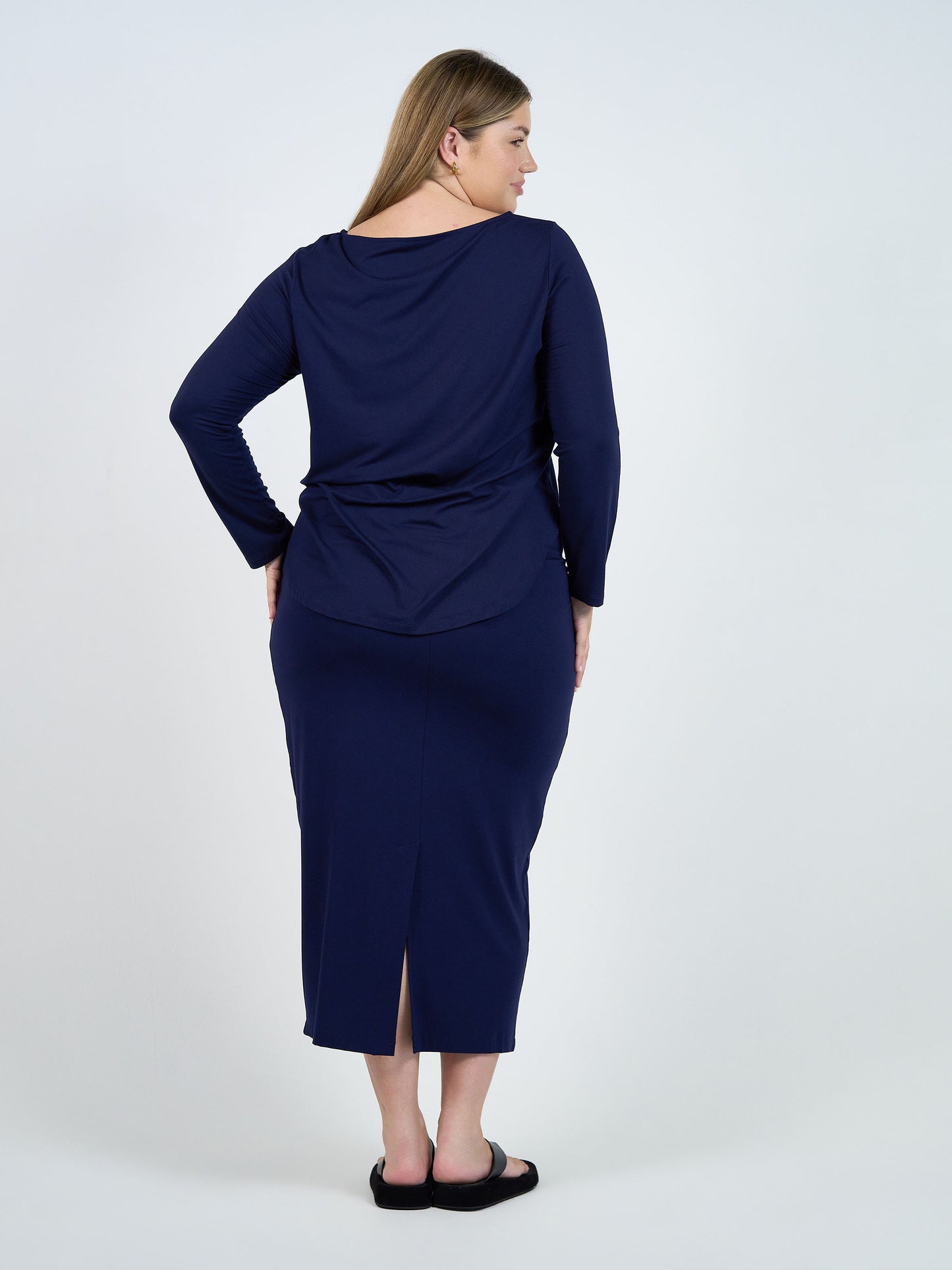 A woman with long blonde hair stands barefoot on a white background, wearing the Maxi Bamboo Whitney Tube Skirt in navy and a matching long-sleeve top, facing away from the camera.