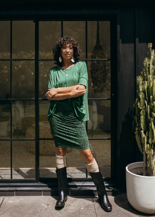 A woman with curly hair in glasses, a green top, the Midi Bamboo Whitney Tube Skirt, black boots, and beige leg warmers stands confidently with crossed arms by a glass door next to a tall potted cactus.