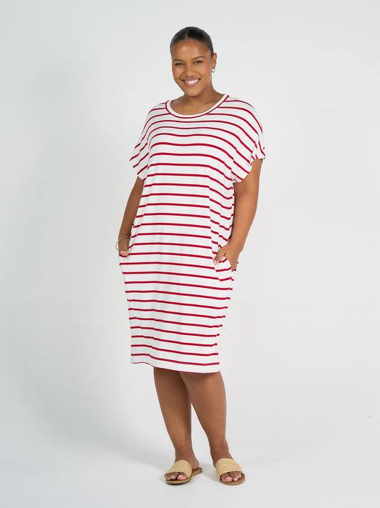 A woman smiles in the Nicks Bamboo Short Sleeve T-Shirt Dress - Lipstick & White Stripe, featuring red and white stripes, short sleeves, pockets, and an oversized fit. She wears tan sandals with her hair pulled back against a white background.