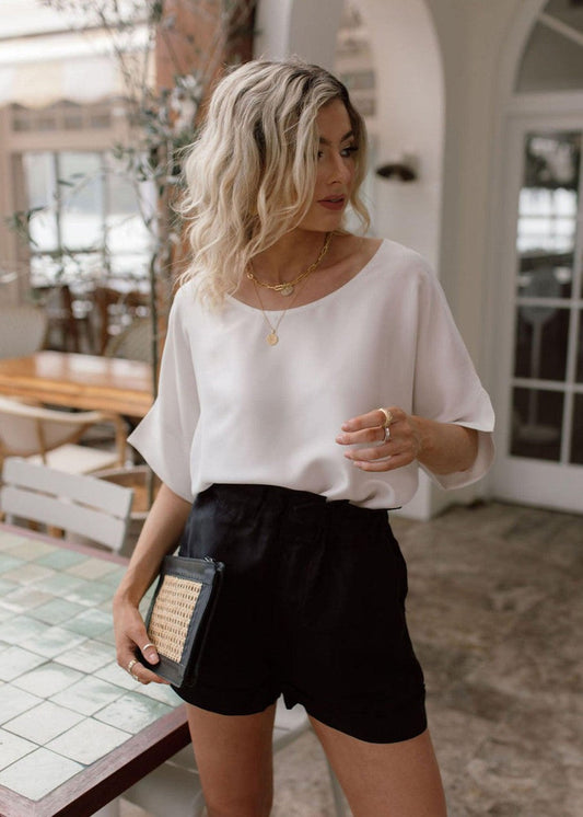 A woman with wavy blonde hair wears the Dua Oversized Silk Top, black high-waisted shorts, layered gold necklaces, and holds a small clutch while standing in a chic indoor-outdoor space with tables and chairs.