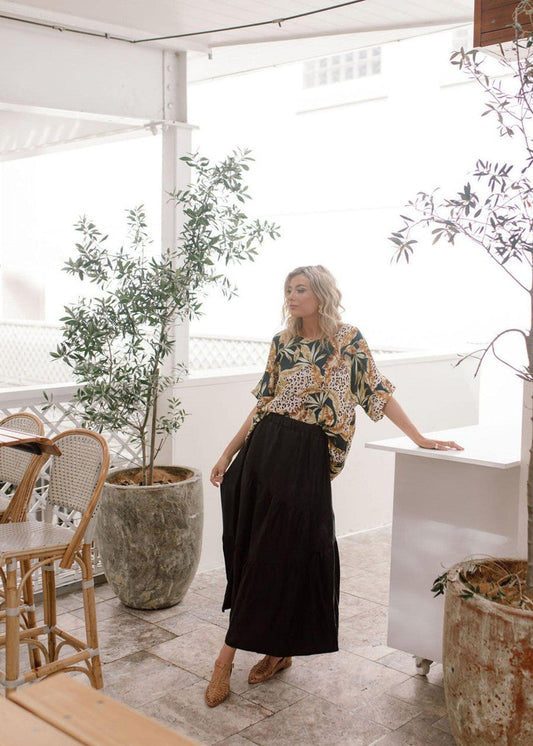 A woman with blonde hair stands in a bright, plant-filled room, wearing a loose blouse and the Summer Silk Maxi Skirt - Black as part of her sustainable fashion look, leaning on a white counter next to wicker chairs.