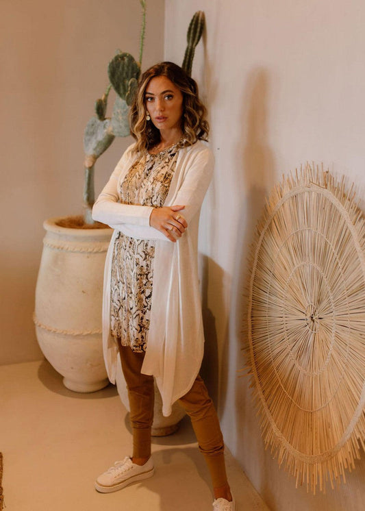 Indoors, a woman stands by a large potted cactus and woven wall art. She wears the Lauper Cashmere/Bamboo Cardigan in Pearl over a patterned dress, with tan pants and white sneakers, arms crossed in a neutral pose.