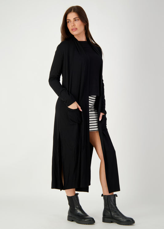 A woman stands confidently with hands in her Bamboo Longline Pocket Cardigan, styled over a black top and striped skirt. Finished with black ankle boots, this sustainable look is perfect for travel and captures effortless style.