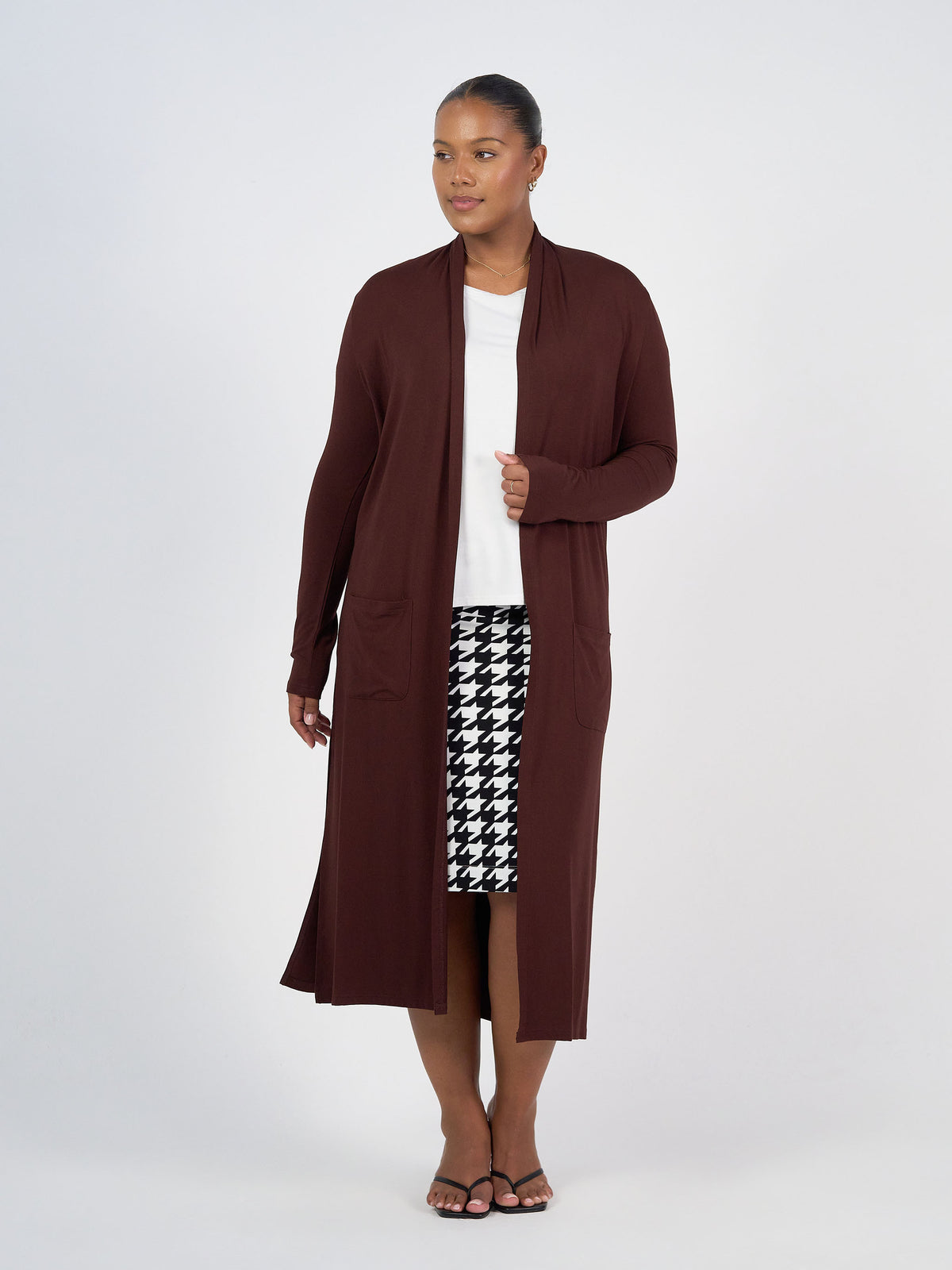 A woman stands against a plain background wearing the Bamboo Longline Pocket Cardigan in Burgundy over a white top and a black-and-white houndstooth skirt with black sandals—a sustainable wardrobe essential.