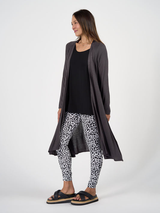 A woman stands sideways, smiling, wearing the Quatro Cashmere/Bamboo Duster Jacket in gray over a black top, with white and black leopard-print leggings and black sandals against a plain white background.