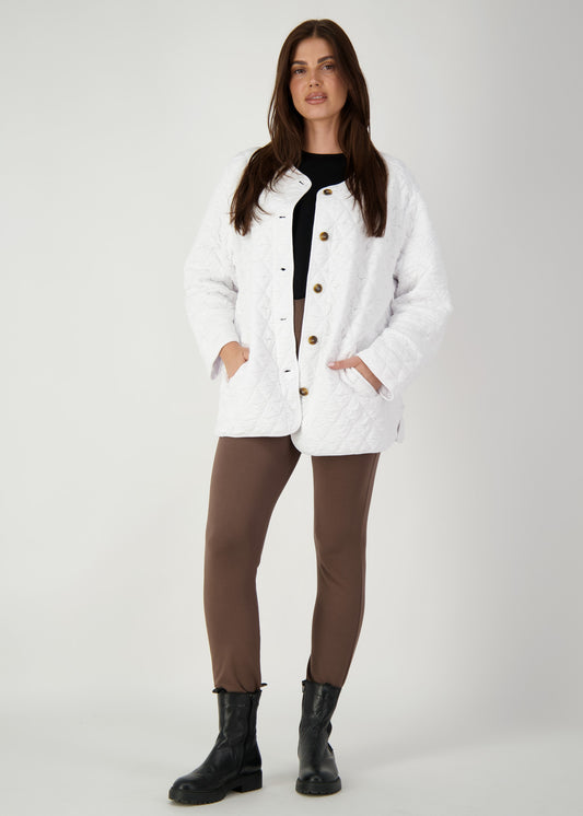 A woman with long brown hair stands against a plain background wearing the Quincy Quilted Bamboo Oversized Jacket, featuring brown buttons, over a black top with brown pants and black boots. Her hands are tucked in the jacket pockets.
