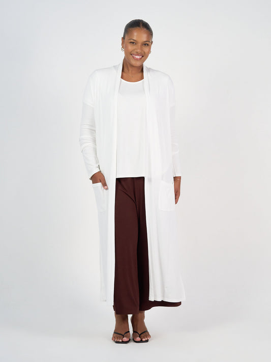 A woman smiles while wearing the Ribbed Bamboo Long-Line Pocket Cardigan over a white top and brown wide-leg pants, standing against a plain white background with her hands in the cardigan’s pockets and black sandals on her feet.