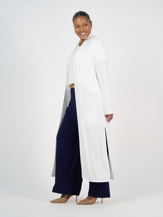 A woman smiles and poses sideways in a Ribbed Bamboo Long-Line Pocket Cardigan worn over a white top, navy wide-leg pants, and beige heels against a plain white background.