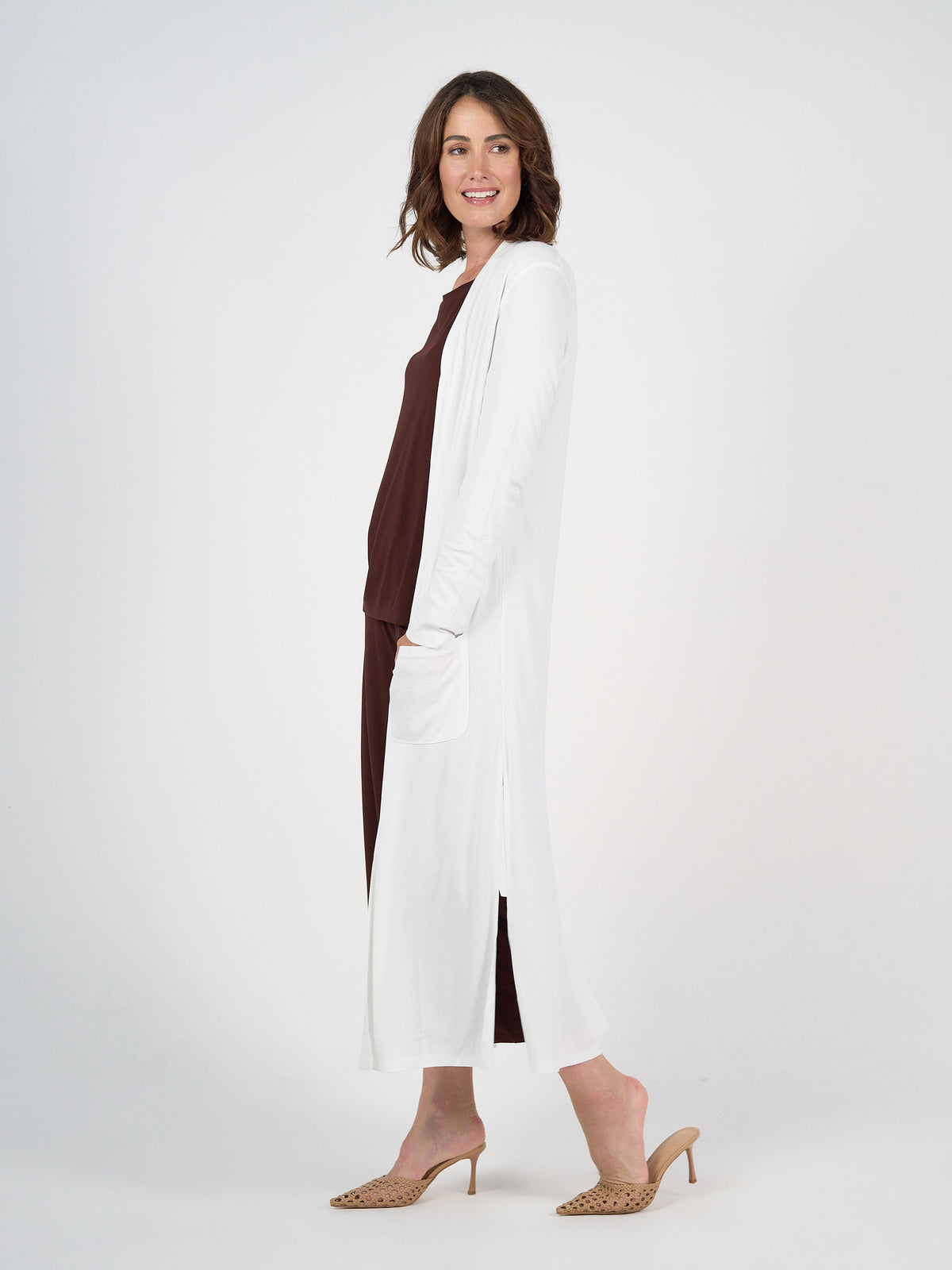 A woman stands sideways, smiling, wearing the Ribbed Bamboo Long-Line Pocket Cardigan over a brown dress with tan heeled mules. The background is plain white.