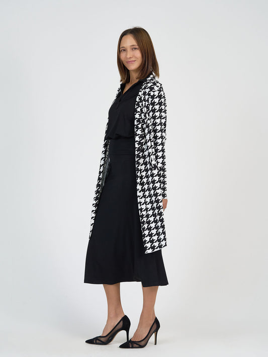 A woman stands smiling in a black blouse and skirt with a long, black-and-white houndstooth patterned coat and black high-heeled shoes against a plain white background.
