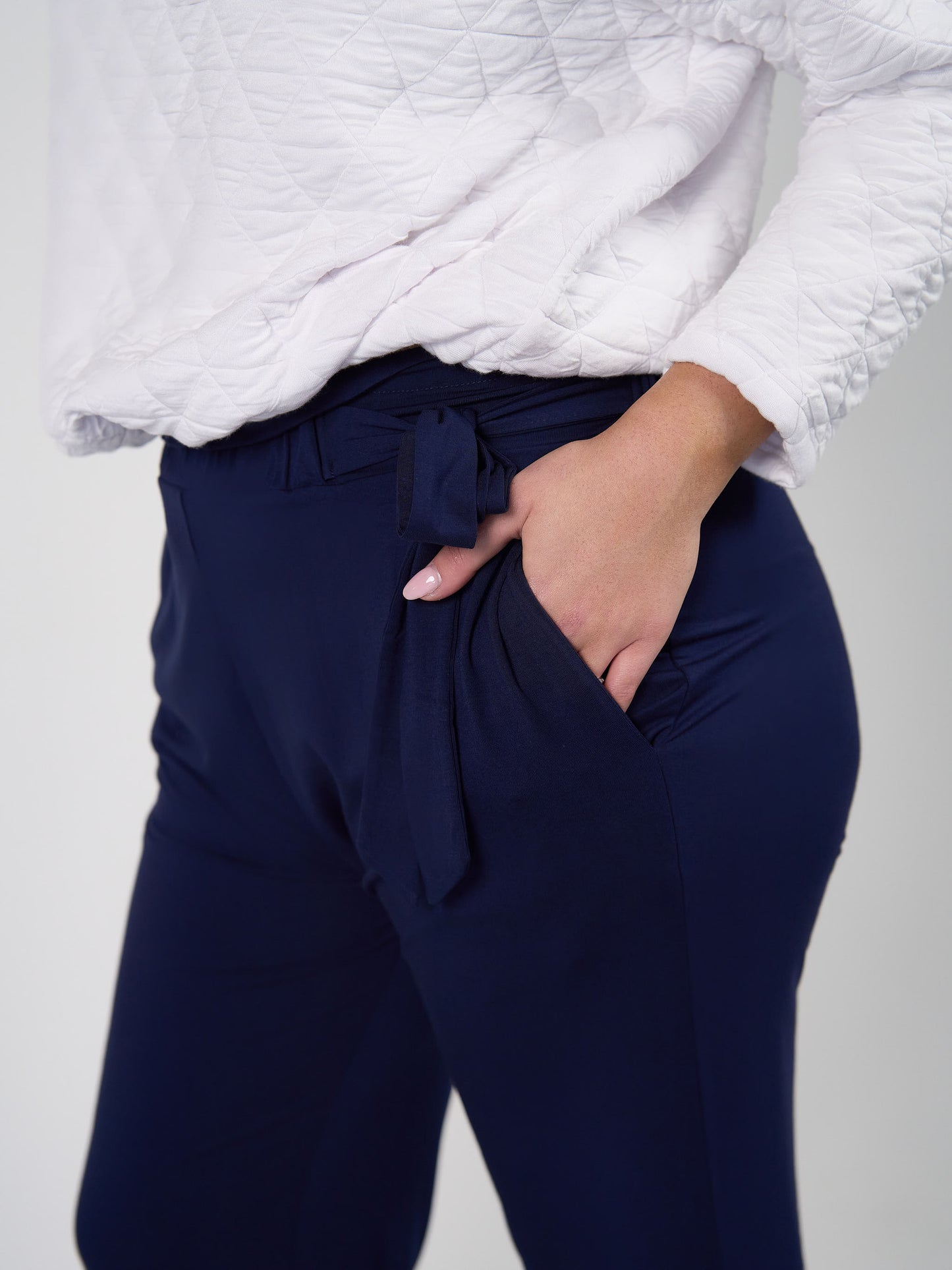 A person wears a textured white top and the Tyler Bamboo Paperbag Pant in navy, featuring a tied waist belt, shown from mid-torso to thigh with one hand in the pant’s pocket.