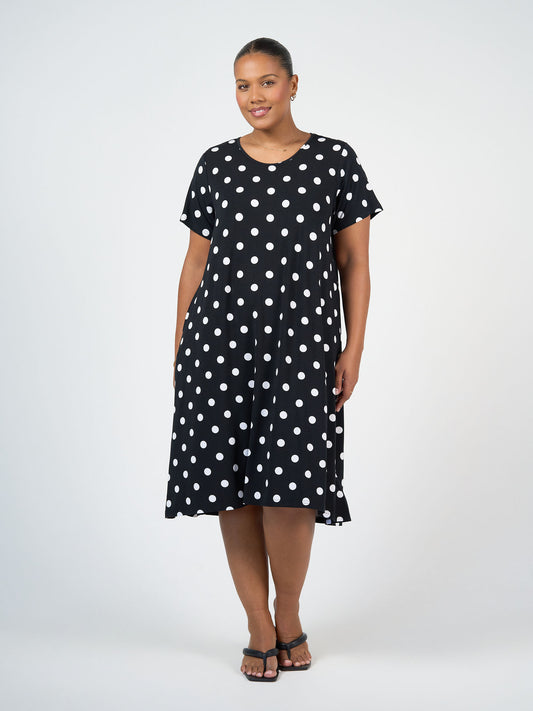 A woman faces forward, smiling slightly, in the Sade Bamboo Oversized T-Shirt Midi Dress - Polka Dot: a knee-length black bamboo jersey dress with white dots, short sleeves, and hidden pockets. She's against a plain light background with black sandals.