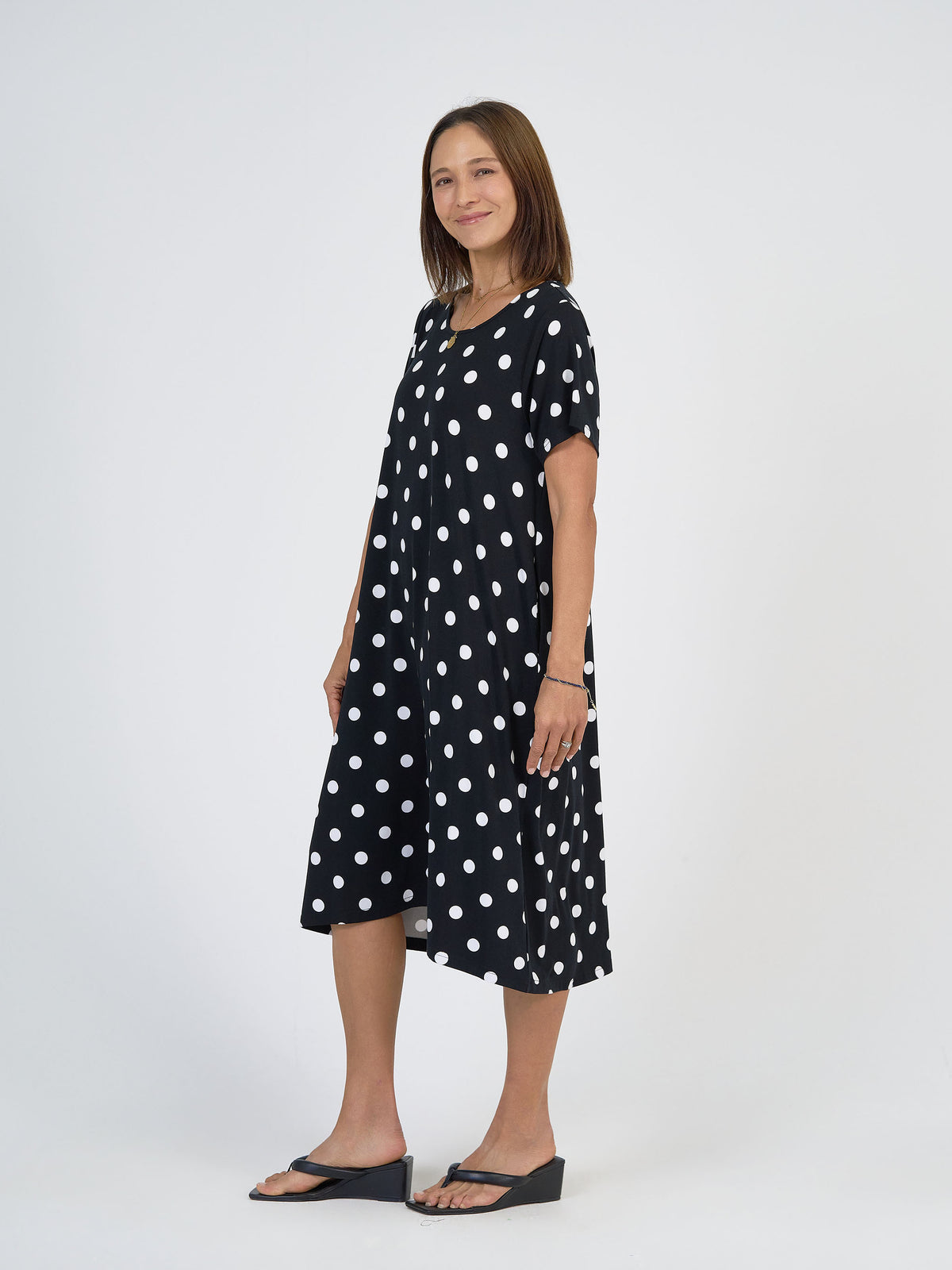 A woman smiles in the Sade Bamboo Oversized T-Shirt Midi Dress - Polka Dot, featuring a loose fit and hidden pockets, paired with black wedge sandals, standing against a plain white background.