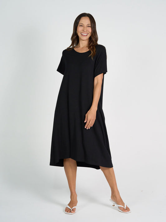 A woman smiles in the Sade Bamboo Oversized T-Shirt Midi Dress in black, a loose-fitting, short-sleeve midi dress with hidden pockets, paired with white heeled sandals against a plain white background.