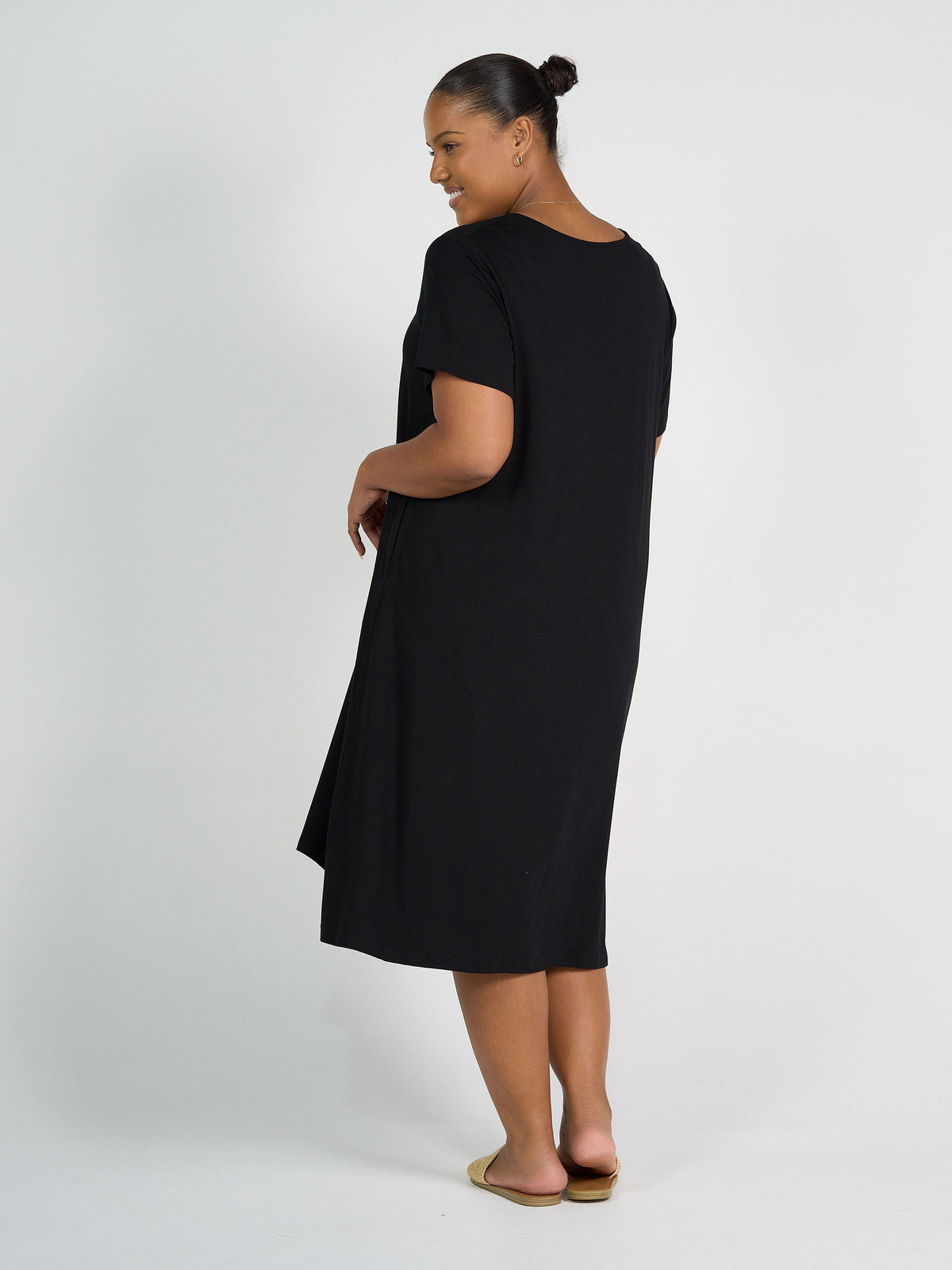 A woman with her hair in a bun smiles at the camera, wearing the Sade Bamboo Oversized T-Shirt Midi Dress - Black with hidden pockets and tan flats, standing sideways against a plain white background.