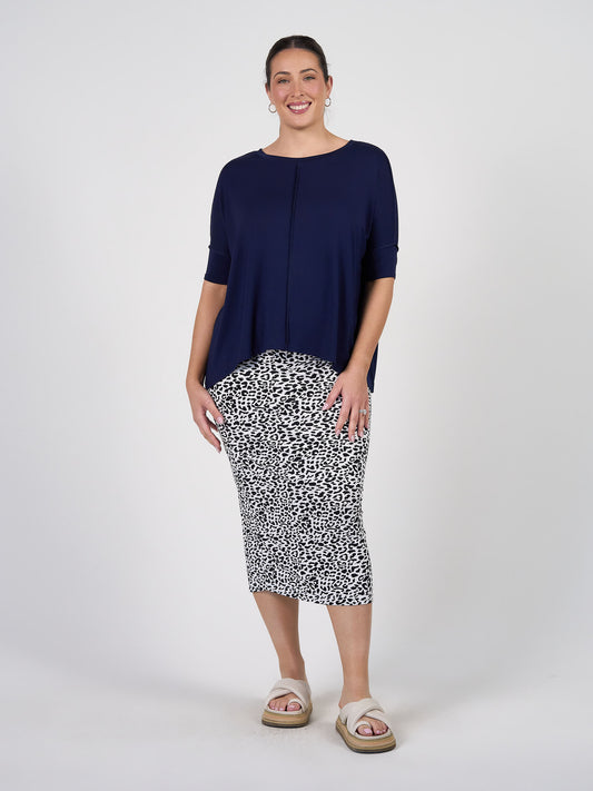 A woman stands smiling against a plain background, wearing a loose navy blue top, the Maxi Bamboo Whitney Tube Skirt in black and white animal print, and beige platform sandals.