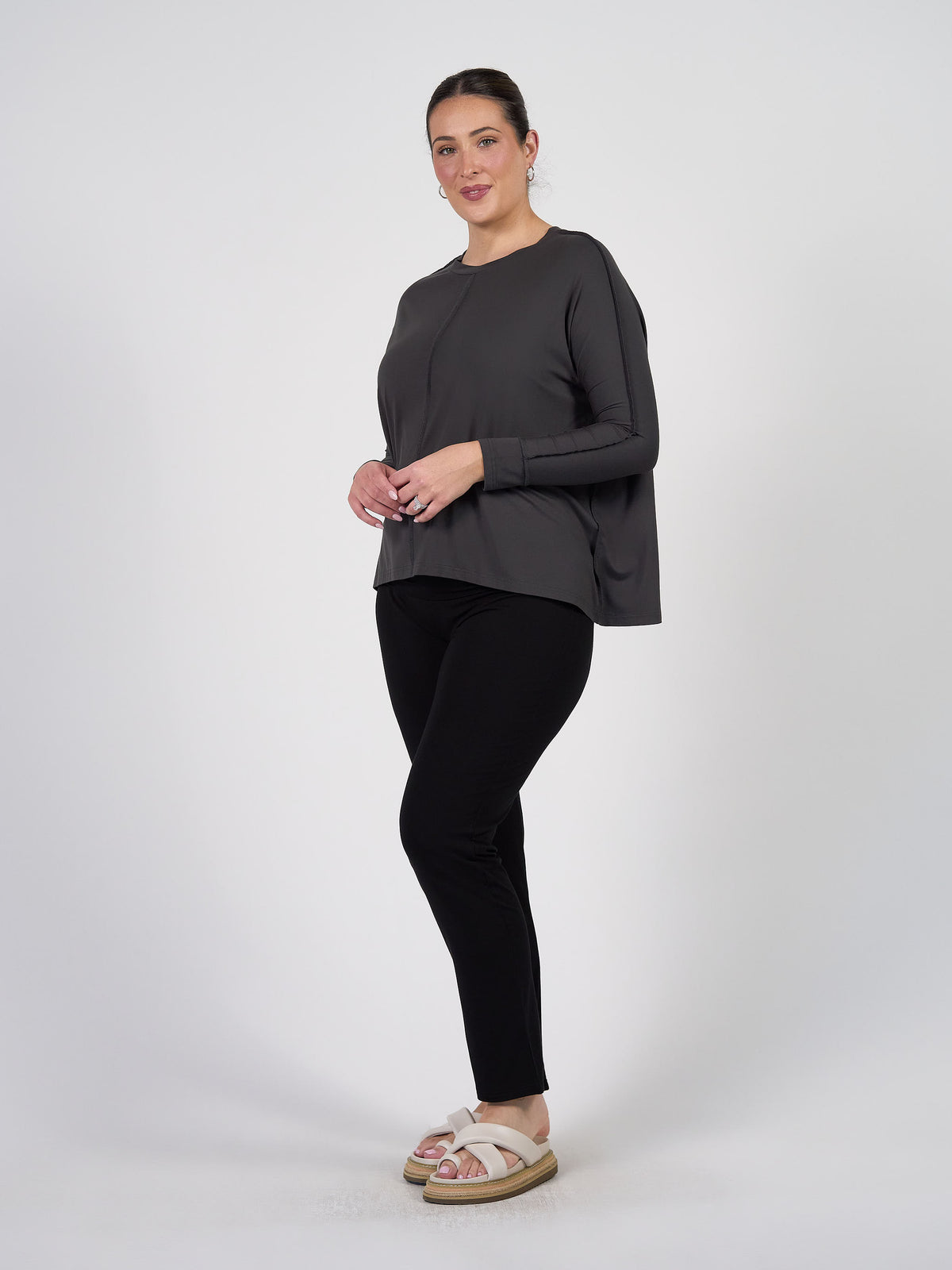 A woman stands against a white background wearing the Stella Longsleeve Oversized Bamboo Slouch Top in Steel, paired with black pants and light-colored sandals. She smiles softly and clasps her hands—an ideal travel outfit.