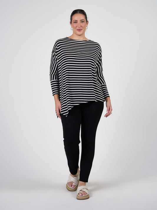 A woman stands against a plain background, smiling slightly with her hair tied back, wearing the Susie Oversized Asymmetrical Bamboo Top in black and white stripe, paired with black pants and white sandals.