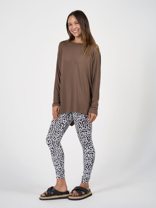 A woman smiles, wearing a loose brown long-sleeve top, Luxe Bamboo Full-Length Legging in black and white leopard print, and black platform slide sandals against a plain white background.