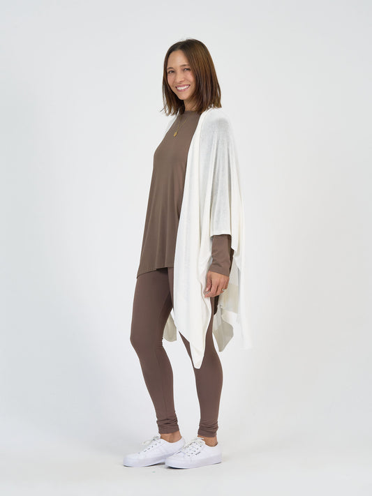 A woman smiles, wearing a taupe long-sleeve top, matching leggings, the Supreme Cashmere/Bamboo Travel Wrap in Cream draped over her shoulders, and white sneakers against a plain white background.