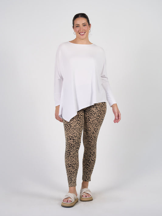 A woman smiles in the Susie Oversized Asymmetrical Bamboo Top - White, styled with leopard print leggings, large hoop earrings, and platform sandals as she poses against a plain white background.