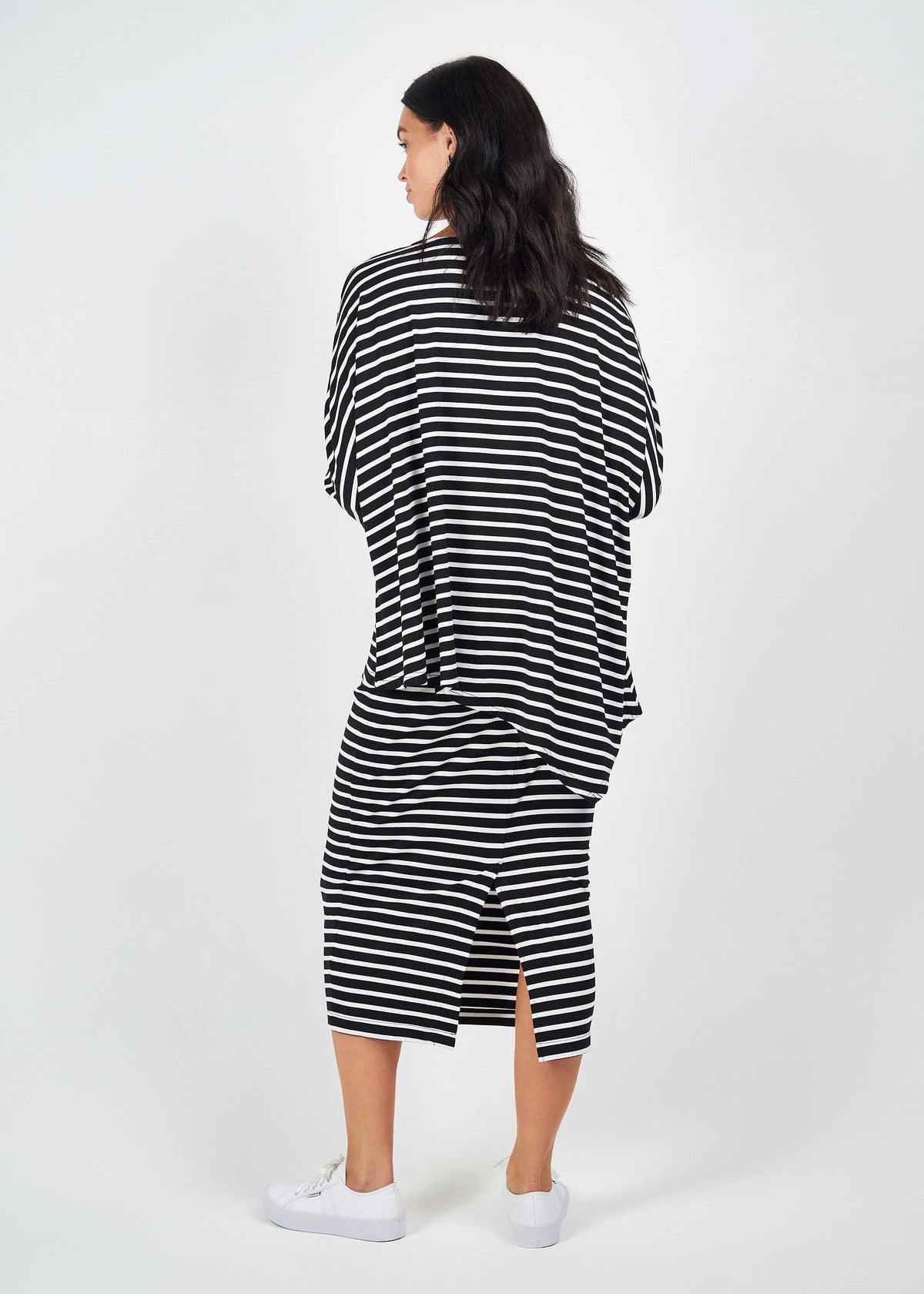 A woman with dark hair faces away, wearing the Susie Oversized Asymmetrical Bamboo Top in black & white stripe and a matching midi skirt with white sneakers, set against a plain white background.