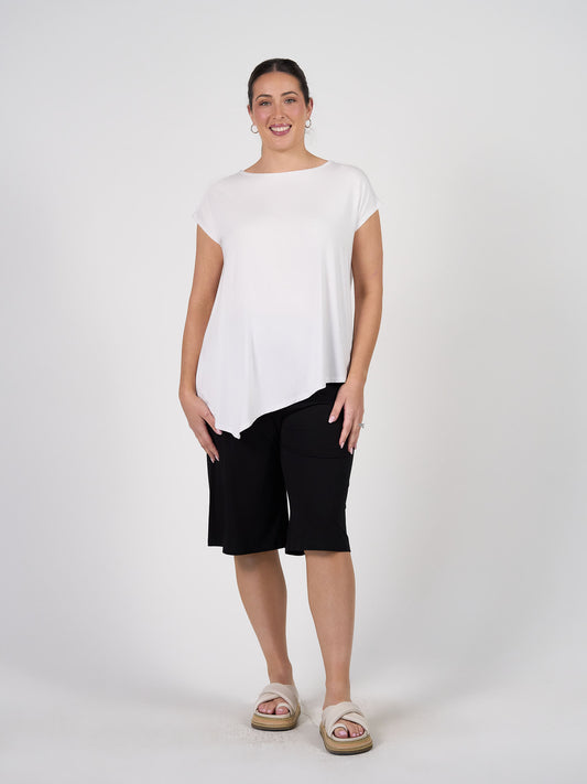 A woman stands against a plain white background, smiling with her hands relaxed at her sides. She wears the Toni Asymmetrical Bamboo T-Shirt in white, black knee-length shorts, white platform sandals, and hoop earrings.