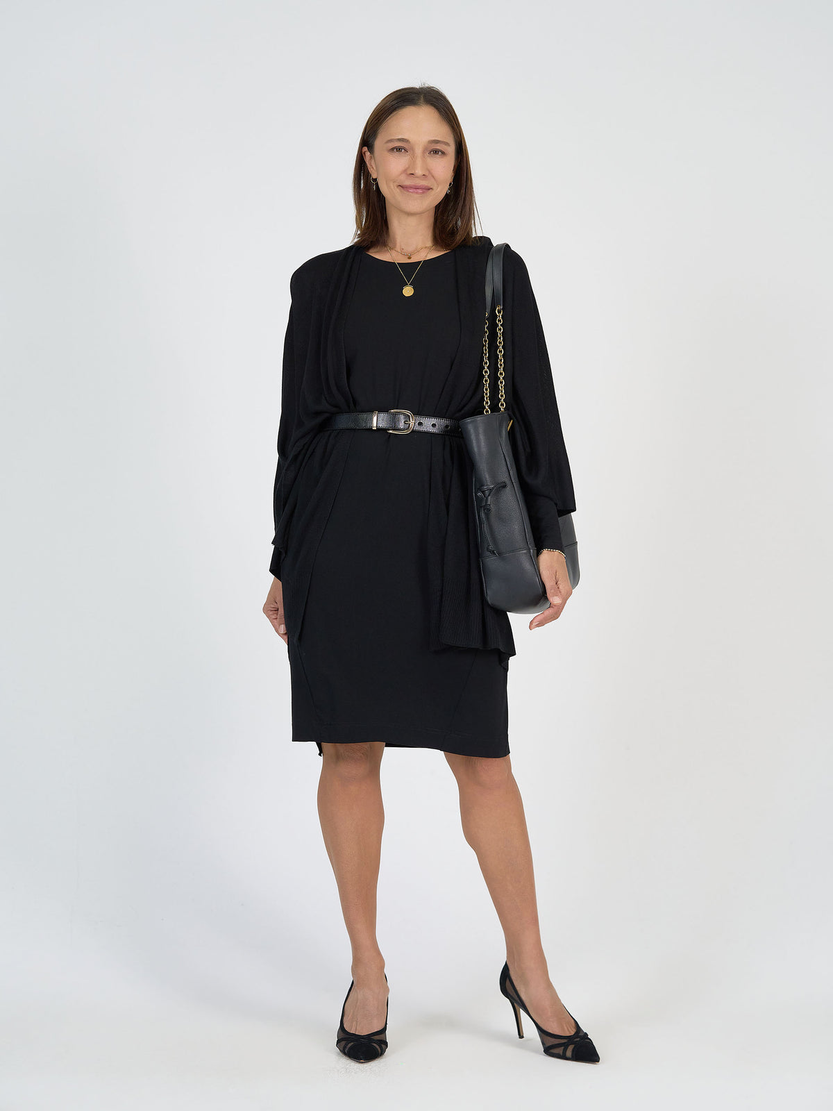 A woman stands against a plain white background, wearing a black knee-length dress with a belt, black high heels, a black shoulder bag, and a gold necklace. She is smiling and looking at the camera.