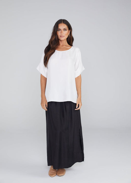 A woman with long brown hair stands facing forward, wearing the Dua Oversized Silk Top in Pearl, a long black skirt, and tan shoes against a plain light gray background.